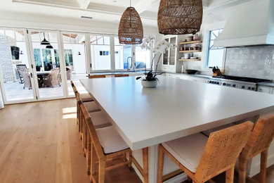 Photo of an expansive nautical u-shaped kitchen in San Diego with a belfast sink, recessed-panel cabinets, white cabinets, engineered stone countertops, white splashback, ceramic splashback, stainless steel appliances, medium hardwood flooring, an island, grey worktops and a coffered ceiling.