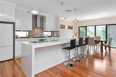 This is an example of a large modern galley eat-in kitchen in Brisbane with a double-bowl sink, flat-panel cabinets, white cabinets, glass sheet splashback, white appliances, medium hardwood floors, with island, quartz benchtops, grey splashback and white benchtop.