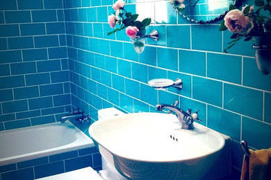 This is an example of a small midcentury 3/4 bathroom in Sydney with a wall-mount sink, furniture-like cabinets, medium wood cabinets, a drop-in tub, a shower/bathtub combo, a two-piece toilet, blue tile, ceramic tile, blue walls and travertine floors.