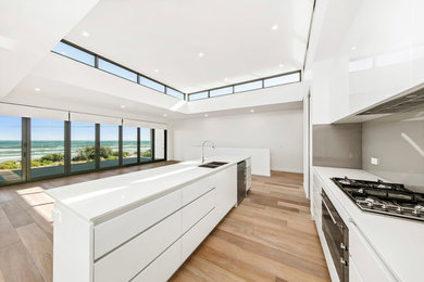 Photo of a contemporary galley kitchen in Melbourne with a double-bowl sink, flat-panel cabinets, white cabinets, brown splashback, black appliances, light hardwood floors, with island, brown floor and white benchtop.