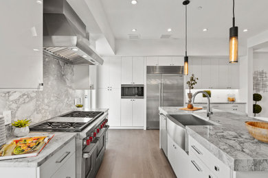Example of a trendy l-shaped medium tone wood floor and brown floor kitchen design in Orlando with a farmhouse sink, flat-panel cabinets, white cabinets, gray backsplash, stainless steel appliances, an island and gray countertops