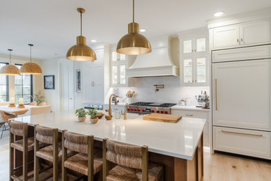 Diseño de cocina comedor lineal grande con fregadero bajoencimera, armarios estilo shaker, puertas de armario blancas, encimera de cuarzo compacto, salpicadero blanco, salpicadero de azulejos tipo metro, electrodomésticos con paneles, suelo de madera clara, una isla, suelo marrón y encimeras blancas