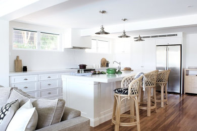 This is an example of a beach style l-shaped open plan kitchen in Sydney with a farmhouse sink, shaker cabinets, white cabinets, granite benchtops, white splashback, ceramic splashback, stainless steel appliances, with island, white benchtop, medium hardwood floors and brown floor.