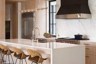 Eat-in kitchen - mid-sized transitional single-wall travertine floor and beige floor eat-in kitchen idea in Austin with a farmhouse sink, flat-panel cabinets, beige cabinets, solid surface countertops, gray backsplash, stone slab backsplash, stainless steel appliances and an island