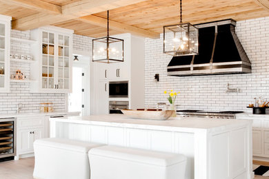 Photo of an expansive transitional l-shaped kitchen in New York with shaker cabinets, white cabinets, white splashback, subway tile splashback, stainless steel appliances, light hardwood floors, with island, beige floor, quartz benchtops and a farmhouse sink.