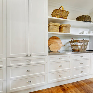 Foto de cuarto de lavado lineal, tradicional, grande, con fregadero integrado, puertas de armario blancas, encimera de acero inoxidable, paredes blancas, suelo de madera en tonos medios y lavadora y secadora apiladas