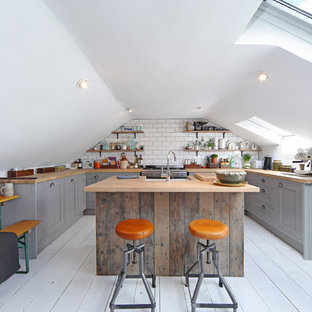 Пример оригинального дизайна интерьера: маленькая п-образная кухня в стиле лофт с обеденным столом, раковиной в стиле кантри, серыми фасадами, столешницей из дерева, техникой из нержавеющей стали, деревянным полом, островом, фасадами с выступающей филенкой, белым фартуком и фартуком из плитки кабанчик