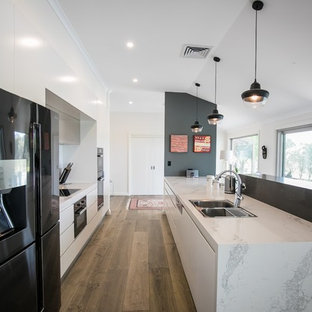 Photo of a contemporary galley kitchen in Sydney with a drop-in sink, flat-panel cabinets, white cabinets, black appliances, medium hardwood floors, a peninsula, brown floor and grey benchtop.