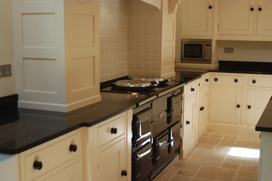 Design ideas for a large traditional u-shaped open plan kitchen in Sussex with a belfast sink, beaded cabinets, granite worktops, beige splashback, ceramic splashback, black appliances, limestone flooring, an island, beige floors and black worktops.