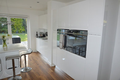 Photo of a large modern galley open plan kitchen in Cheshire with a single-bowl sink, flat-panel cabinets, white cabinets, composite countertops, metallic splashback, mirror splashback, coloured appliances, medium hardwood flooring, an island and brown floors.