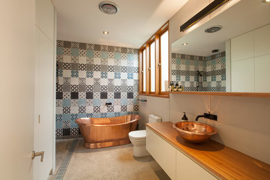 Photo of a mid-sized contemporary master bathroom in Sydney with white cabinets, wood benchtops, multi-coloured tile, a freestanding tub, a one-piece toilet, cement tile, blue walls, concrete floors and a vessel sink.
