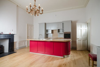 Design ideas for a mid-sized traditional galley open plan kitchen in Other with a farmhouse sink, shaker cabinets, red cabinets, wood benchtops, grey splashback, glass sheet splashback, light hardwood floors and with island.