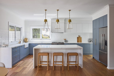 This is an example of a large beach style u-shaped eat-in kitchen in Sydney with a farmhouse sink, shaker cabinets, blue cabinets, quartz benchtops, white splashback, engineered quartz splashback, stainless steel appliances, dark hardwood floors, with island, brown floor and white benchtop.