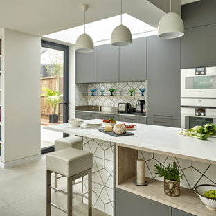 Einzeilige Moderne Wohnküche mit flächenbündigen Schrankfronten, grauen Schränken, Mineralwerkstoff-Arbeitsplatte, Küchenrückwand in Weiß, Rückwand aus Keramikfliesen, Küchengeräten aus Edelstahl, Keramikboden, Kücheninsel und grauem Boden in London