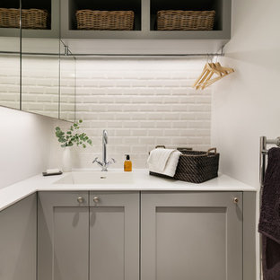 This is an example of a small utility room in London with a built-in sink, recessed-panel cabinets, grey cabinets, composite countertops, white walls, light hardwood flooring and a concealed washer and dryer.