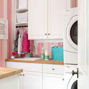 Photo of a classic galley separated utility room in Los Angeles with a built-in sink, raised-panel cabinets, white cabinets, wood worktops, pink walls, a stacked washer and dryer and pink floors.
