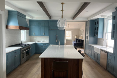 Inspiration for a large beach style u-shaped kitchen in Philadelphia with a farmhouse sink, shaker cabinets, quartz benchtops, white splashback, porcelain splashback, stainless steel appliances, light hardwood floors, with island, white benchtop and exposed beam.