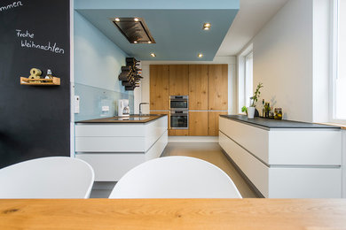Mid-sized contemporary galley open plan kitchen in Berlin with an undermount sink, raised-panel cabinets, white cabinets, solid surface benchtops, blue splashback, glass sheet splashback, stainless steel appliances and concrete floors.
