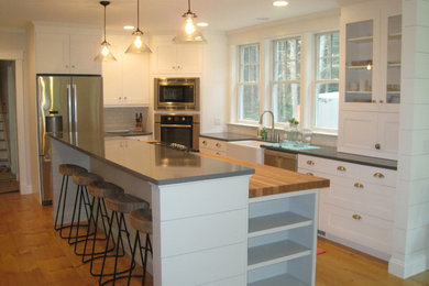 Example of a large transitional u-shaped medium tone wood floor and brown floor kitchen design in Portland Maine with a farmhouse sink, shaker cabinets, white cabinets, white backsplash, subway tile backsplash, stainless steel appliances, an island and black countertops