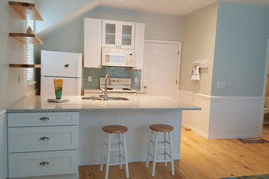 Photo of a mid-sized beach style galley open plan kitchen in Orlando with a double-bowl sink, shaker cabinets, white cabinets, quartz benchtops, grey splashback, subway tile splashback, white appliances, light hardwood floors, a peninsula and beige floor.