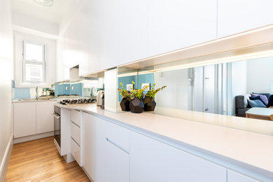 Inspiration for a small contemporary single-wall open plan kitchen in Sydney with a drop-in sink, flat-panel cabinets, white cabinets, quartz benchtops, metallic splashback, mirror splashback, stainless steel appliances, light hardwood floors, brown floor and grey benchtop.