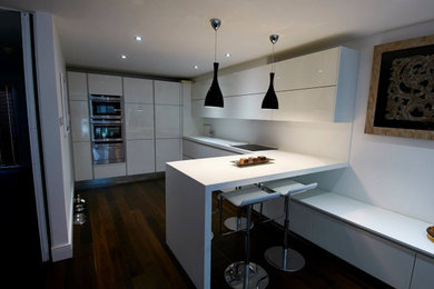 Medium sized contemporary l-shaped open plan kitchen in West Midlands with a built-in sink, flat-panel cabinets, white cabinets, composite countertops, white splashback, stainless steel appliances, dark hardwood flooring, a breakfast bar, brown floors and white worktops.