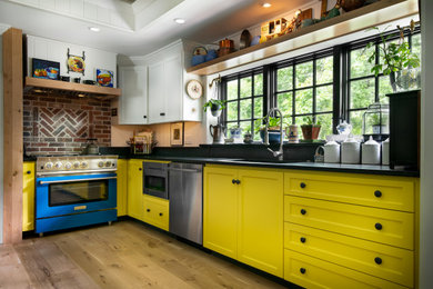 Example of a mid-sized transitional l-shaped light wood floor, brown floor and tray ceiling open concept kitchen design in Kansas City with an undermount sink, shaker cabinets, yellow cabinets, soapstone countertops, black backsplash, wood backsplash, colored appliances, no island and black countertops