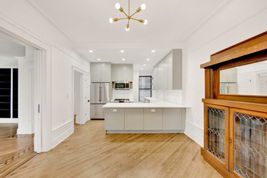 Example of a large transitional l-shaped medium tone wood floor and brown floor eat-in kitchen design in New York with a drop-in sink, flat-panel cabinets, green cabinets, quartz countertops, white backsplash, ceramic backsplash, stainless steel appliances, a peninsula and white countertops