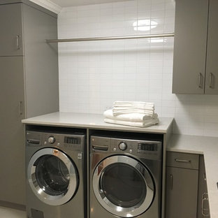 他の地域の大きいトランジショナルスタイルのおしゃれな洗濯室 (ドロップインシンク、フラットパネル扉のキャビネット、グレーのキャビネット、人工大理石カウンター、グレーの壁、磁器タイルの床、左右配置の洗濯機・乾燥機、白い床) の写真