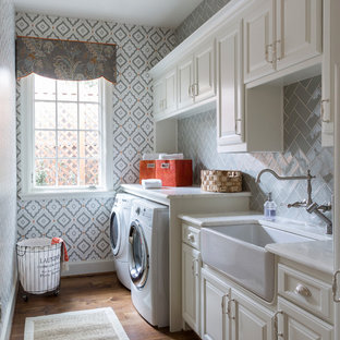 Photo of a classic single-wall separated utility room in Dallas with a belfast sink, raised-panel cabinets, white cabinets, multi-coloured walls, medium hardwood flooring, a side by side washer and dryer, brown floors and white worktops.