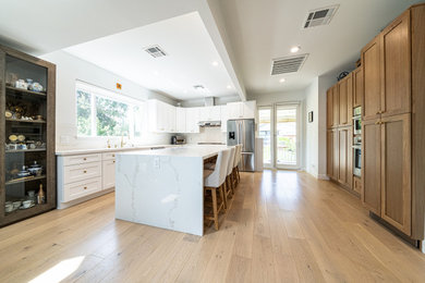 This is an example of a large traditional u-shaped kitchen/diner in Los Angeles with a submerged sink, shaker cabinets, white cabinets, marble worktops, white splashback, marble splashback, stainless steel appliances, medium hardwood flooring, an island, brown floors, white worktops and exposed beams.