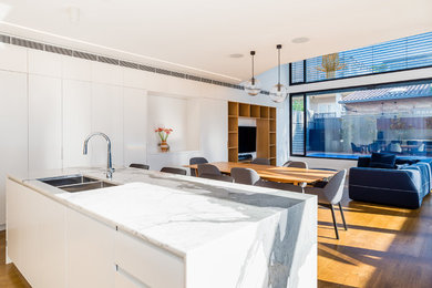Large contemporary open plan kitchen in Sydney with an undermount sink, beaded inset cabinets, white cabinets, marble benchtops, white splashback, porcelain splashback, black appliances, light hardwood floors, with island and brown floor.