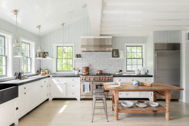 Design ideas for a beach style l-shaped kitchen in New York with a farmhouse sink, shaker cabinets, white cabinets, grey splashback, subway tile splashback, stainless steel appliances, light hardwood floors, with island, beige floor and black benchtop.