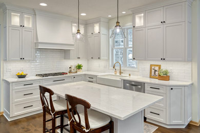 Mid-sized transitional u-shaped medium tone wood floor and brown floor eat-in kitchen photo in Minneapolis with a farmhouse sink, recessed-panel cabinets, white cabinets, quartz countertops, white backsplash, ceramic backsplash, stainless steel appliances, an island and white countertops