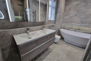 Inspiration for a large contemporary grey and brown ensuite bathroom in West Midlands with flat-panel cabinets, grey cabinets, a freestanding bath, a built-in shower, a wall mounted toilet, grey tiles, porcelain tiles, grey walls, porcelain flooring, a wall-mounted sink, grey floors, a hinged door, a wall niche, double sinks and a floating vanity unit.