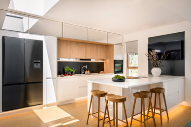 Photo of a large contemporary open plan kitchen in Hampshire with an integrated sink, flat-panel cabinets, white cabinets, terrazzo worktops, metallic splashback, metal splashback, black appliances, light hardwood flooring, an island, brown floors and white worktops.