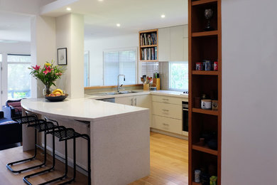 This is an example of a mid-sized contemporary u-shaped eat-in kitchen in Melbourne with a drop-in sink, flat-panel cabinets, quartz benchtops, stainless steel appliances, medium hardwood floors, a peninsula, white cabinets and window splashback.