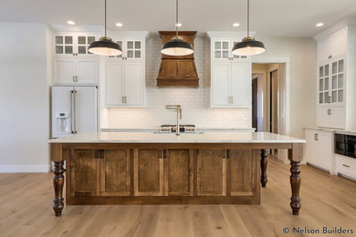 Design ideas for a mid-sized country l-shaped open plan kitchen in Chicago with a farmhouse sink, shaker cabinets, white cabinets, quartz benchtops, white splashback, subway tile splashback, white appliances, light hardwood floors, with island, brown floor and white benchtop.