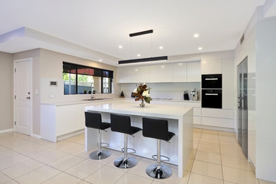 This is an example of a large modern u-shaped separate kitchen in Sydney with a drop-in sink, flat-panel cabinets, white cabinets, quartz benchtops, white splashback, stone slab splashback, black appliances, porcelain floors, with island, beige floor and white benchtop.