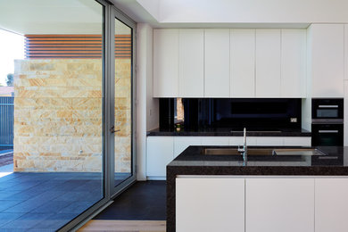 Inspiration for a large contemporary galley eat-in kitchen in Adelaide with an undermount sink, recessed-panel cabinets, white cabinets, granite benchtops, black splashback, glass sheet splashback, black appliances, light hardwood floors and with island.