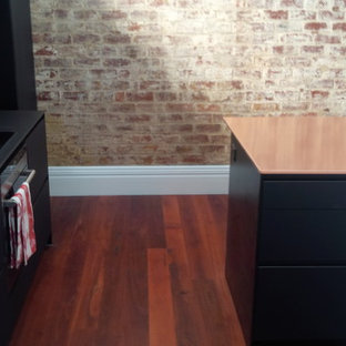 Industriell inredning av ett mellanstort linjärt skafferi, med en enkel diskho, svarta skåp, bänkskiva i akrylsten, stänkskydd med metallisk yta, glaspanel som stänkskydd, svarta vitvaror, mellanmörkt trägolv och en köksö