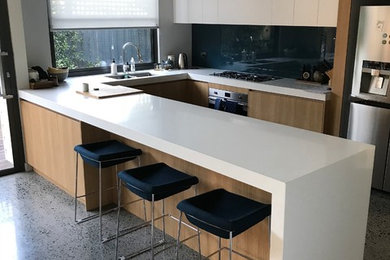 This is an example of a large contemporary u-shaped open plan kitchen in Melbourne with a double-bowl sink, louvered cabinets, light wood cabinets, solid surface benchtops, blue splashback, glass sheet splashback, stainless steel appliances, concrete floors, with island, multi-coloured floor and white benchtop.