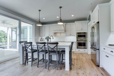 Example of a large beach style light wood floor kitchen design in Baltimore with a farmhouse sink, recessed-panel cabinets, white cabinets, quartz countertops, stainless steel appliances and an island