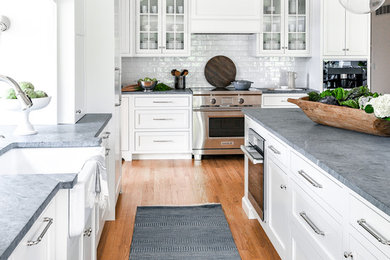 Kitchen - large transitional medium tone wood floor and brown floor kitchen idea in Philadelphia with a farmhouse sink, white cabinets, soapstone countertops, white backsplash, ceramic backsplash, paneled appliances, an island, gray countertops and shaker cabinets