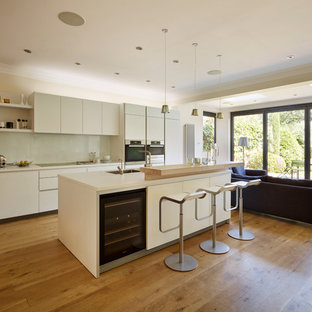 Photo of a contemporary galley open plan kitchen in Cheshire with a submerged sink, flat-panel cabinets, white cabinets, white splashback, glass sheet splashback, medium hardwood flooring, an island and stainless steel appliances.