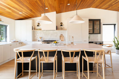 Example of a large minimalist light wood floor and vaulted ceiling kitchen design in Vancouver with a farmhouse sink, shaker cabinets, white cabinets, white backsplash, an island and white countertops