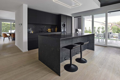 Inspiration for a medium sized modern grey and black galley kitchen in London with a single-bowl sink, flat-panel cabinets, black cabinets, black splashback, black appliances, light hardwood flooring, an island, black worktops and a drop ceiling.