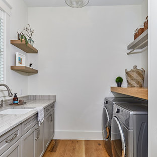 チャールストンの大きいビーチスタイルのおしゃれな洗濯室 (アンダーカウンターシンク、落し込みパネル扉のキャビネット、グレーのキャビネット、白い壁、淡色無垢フローリング、左右配置の洗濯機・乾燥機、グレーのキッチンカウンター、珪岩カウンター) の写真