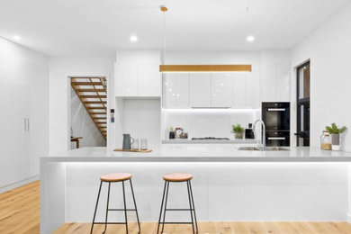 Design ideas for a mid-sized modern galley kitchen pantry in Melbourne with a double-bowl sink, flat-panel cabinets, white cabinets, quartz benchtops, white splashback, glass sheet splashback, black appliances, light hardwood floors, with island, brown floor and grey benchtop.