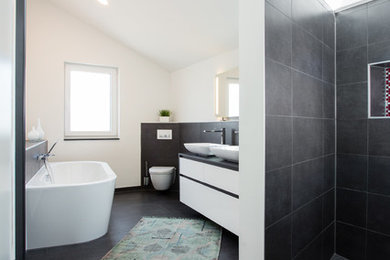 Großes Modernes Badezimmer En Suite mit flächenbündigen Schrankfronten, weißen Schränken, freistehender Badewanne, bodengleicher Dusche, Wandtoilette, schwarzen Fliesen, Keramikfliesen, weißer Wandfarbe, Porzellan-Bodenfliesen, Aufsatzwaschbecken, Waschtisch aus Holz, schwarzem Boden und schwarzer Waschtischplatte in Köln